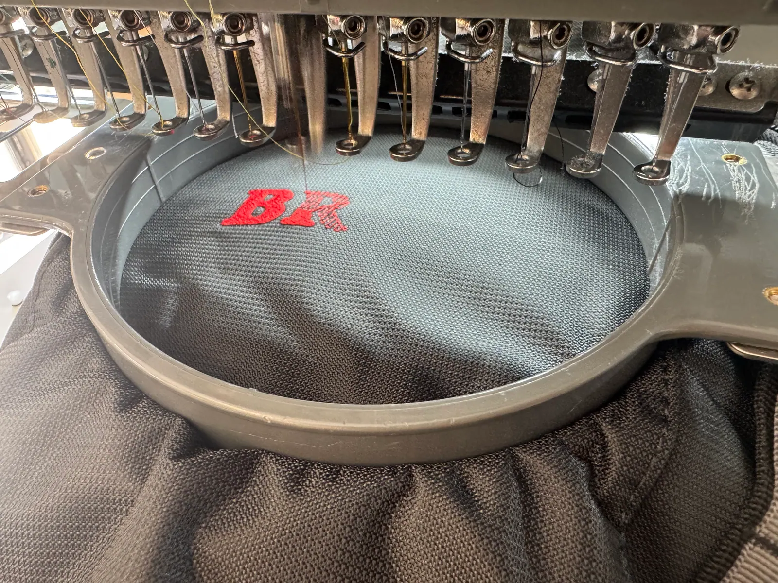 Multi-needle embroidery machine stitching red lettering onto black mesh fabric held in a hoop