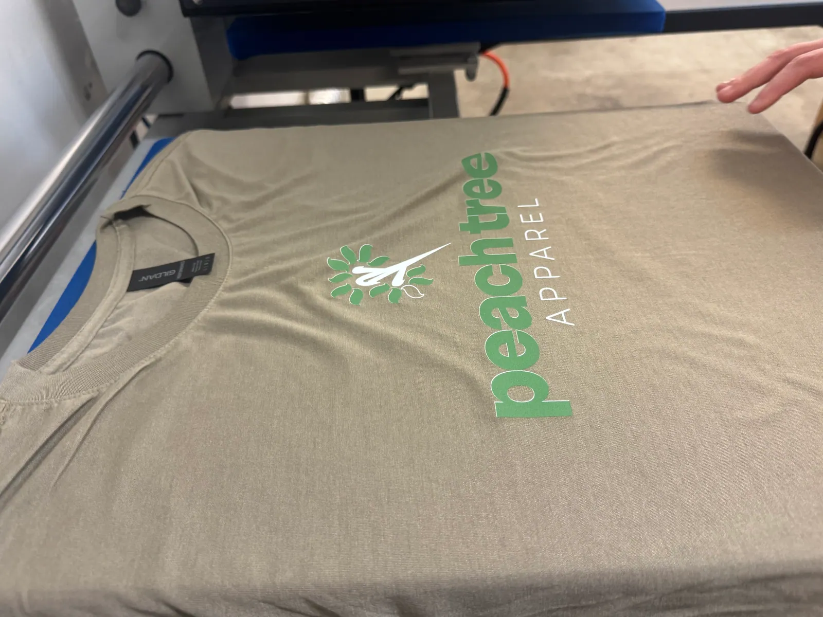 Olive T-shirt with a green and white PeachTree Apparel logo on the front, on the heat press