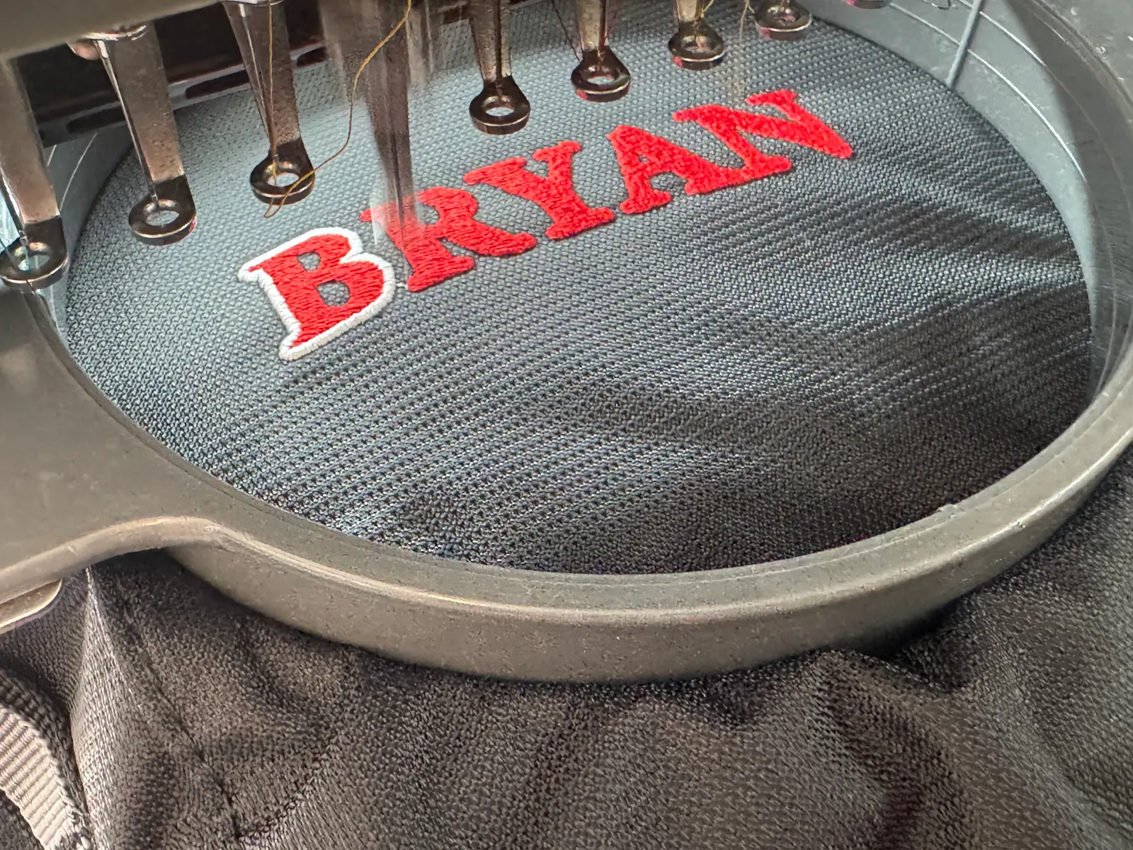 Embroidery needles stitching the name BRYAN in red thread on black mesh held in the hoop